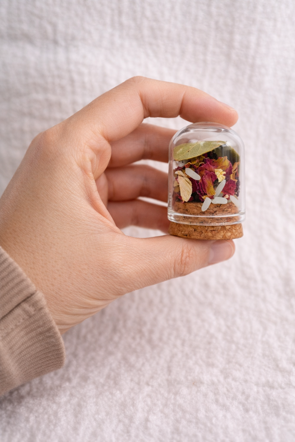 Hand holding a small glass jar with dried herbs against a light background