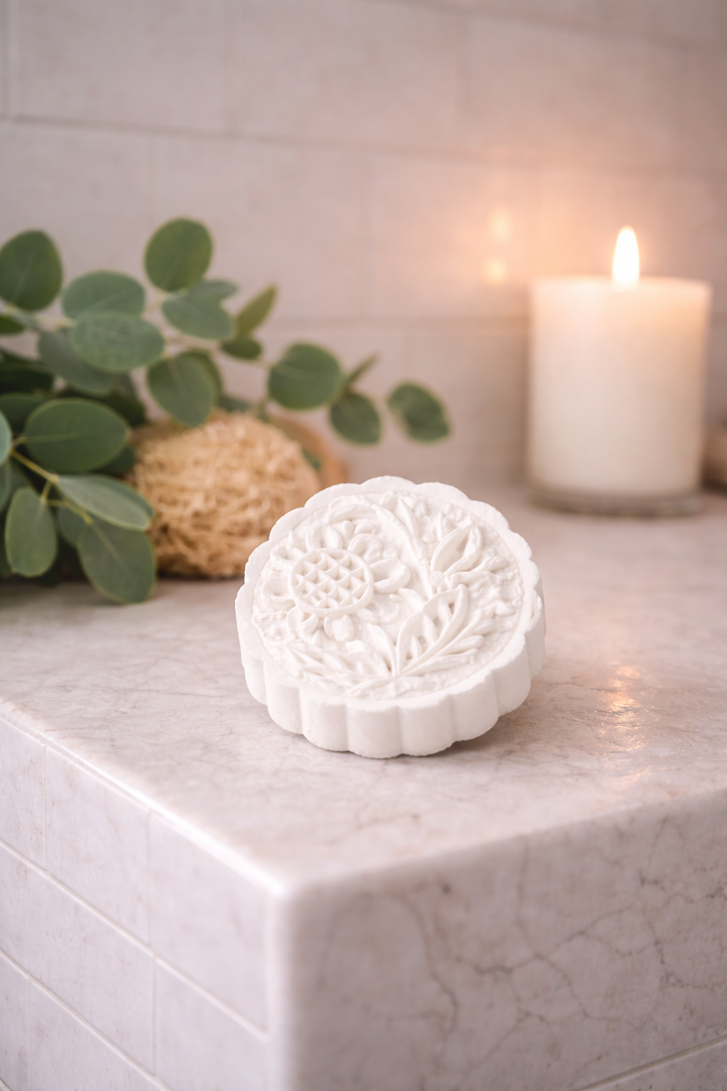 Decorative white mooncake-shaped shower Steamer on a marble surface with a candle and greenery in the background.